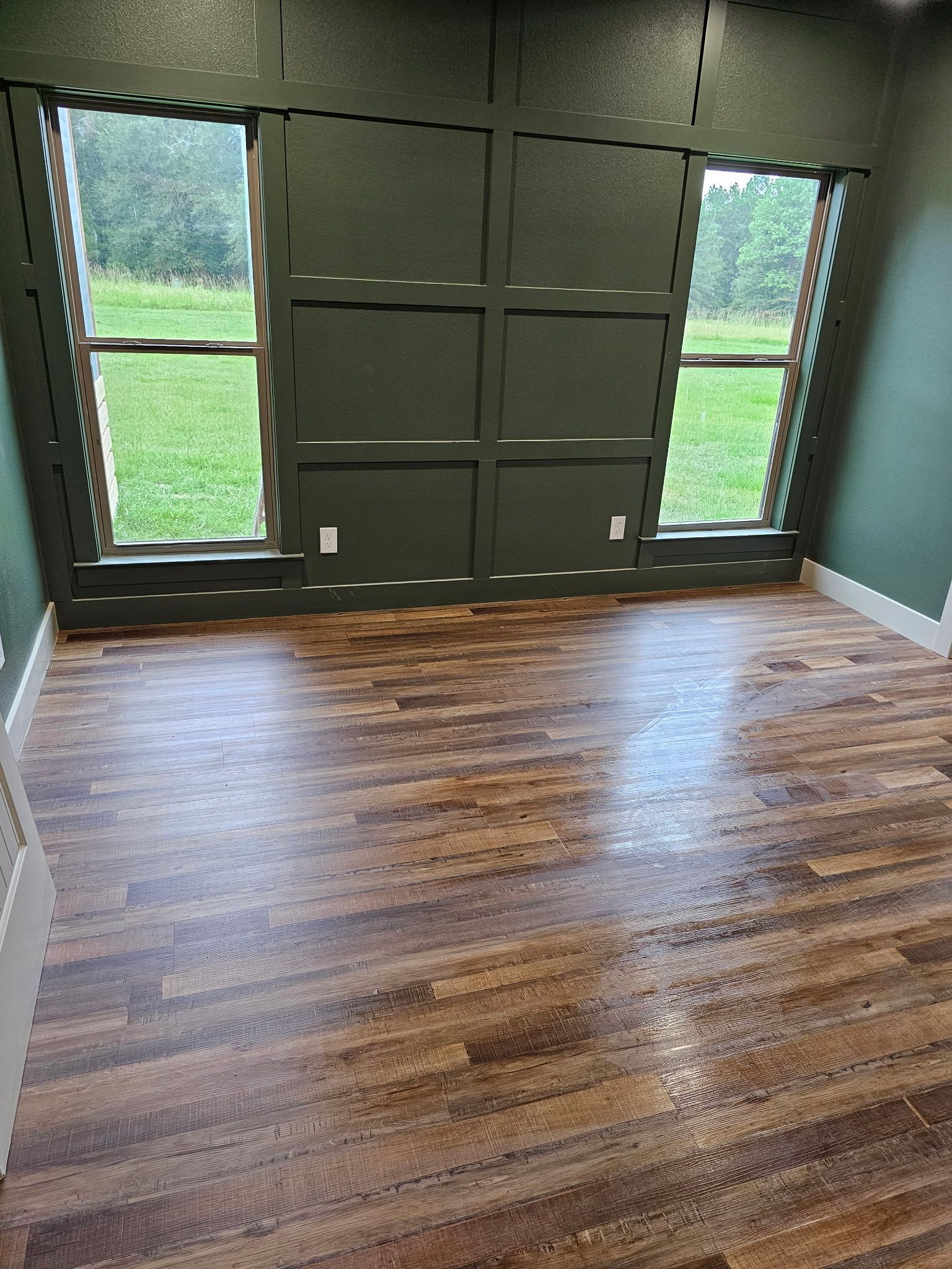 Empty room with dark green paneled walls, two windows looking onto a grassy field, and dark wood-look flooring.