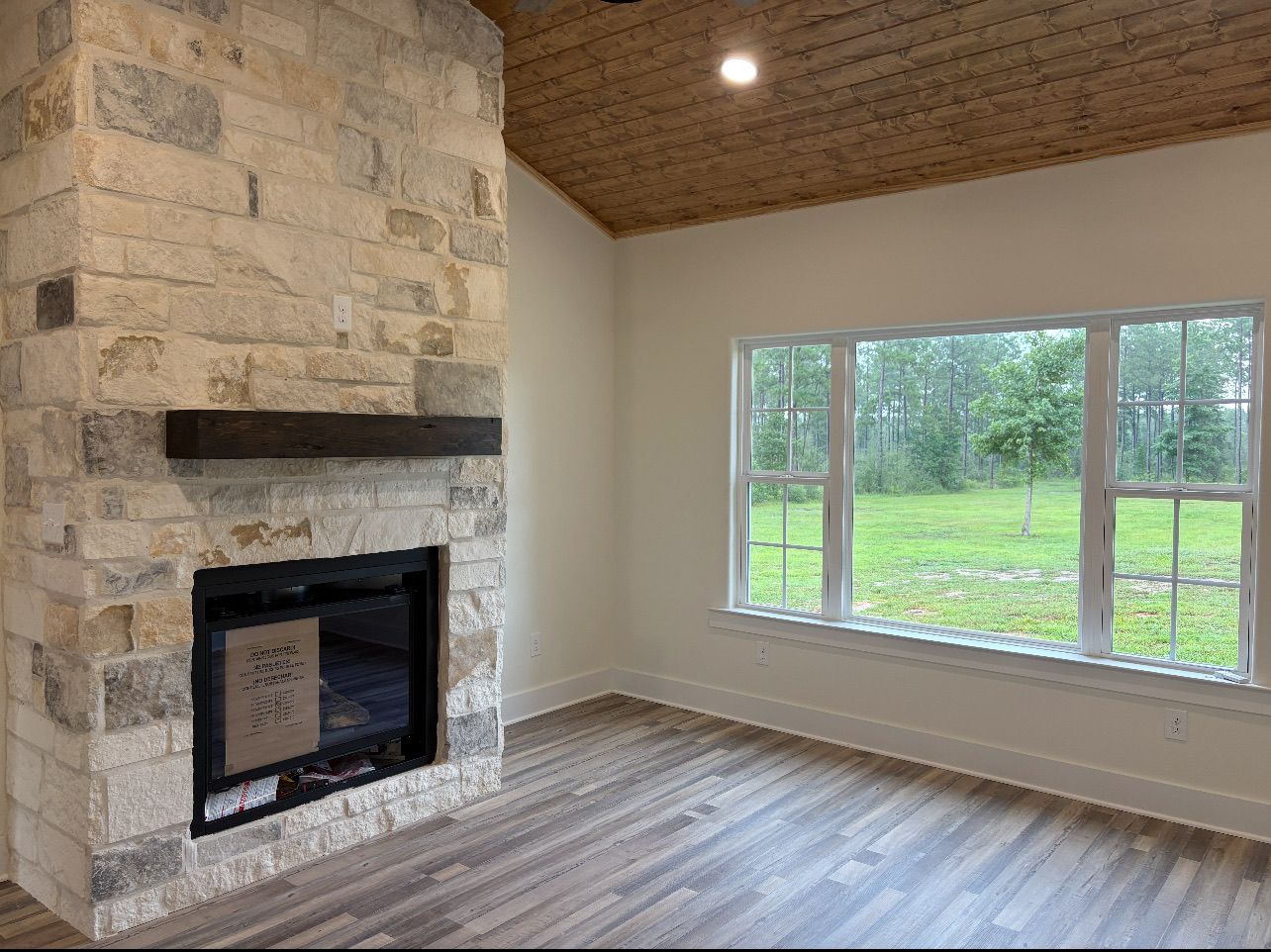 A room featuring a large stone fireplace with a wooden mantel, wood-plank ceiling, and a wide window looking outside.