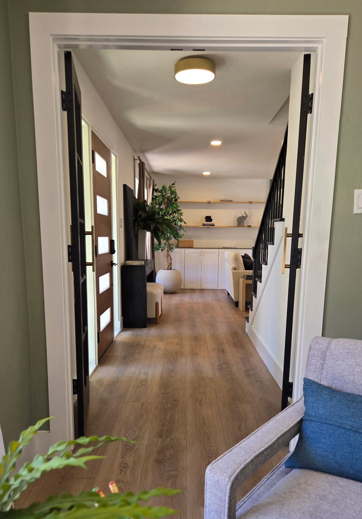 View through a wide doorway into a brightly lit hallway with wood-look flooring, a modern staircase, and floating shelves.