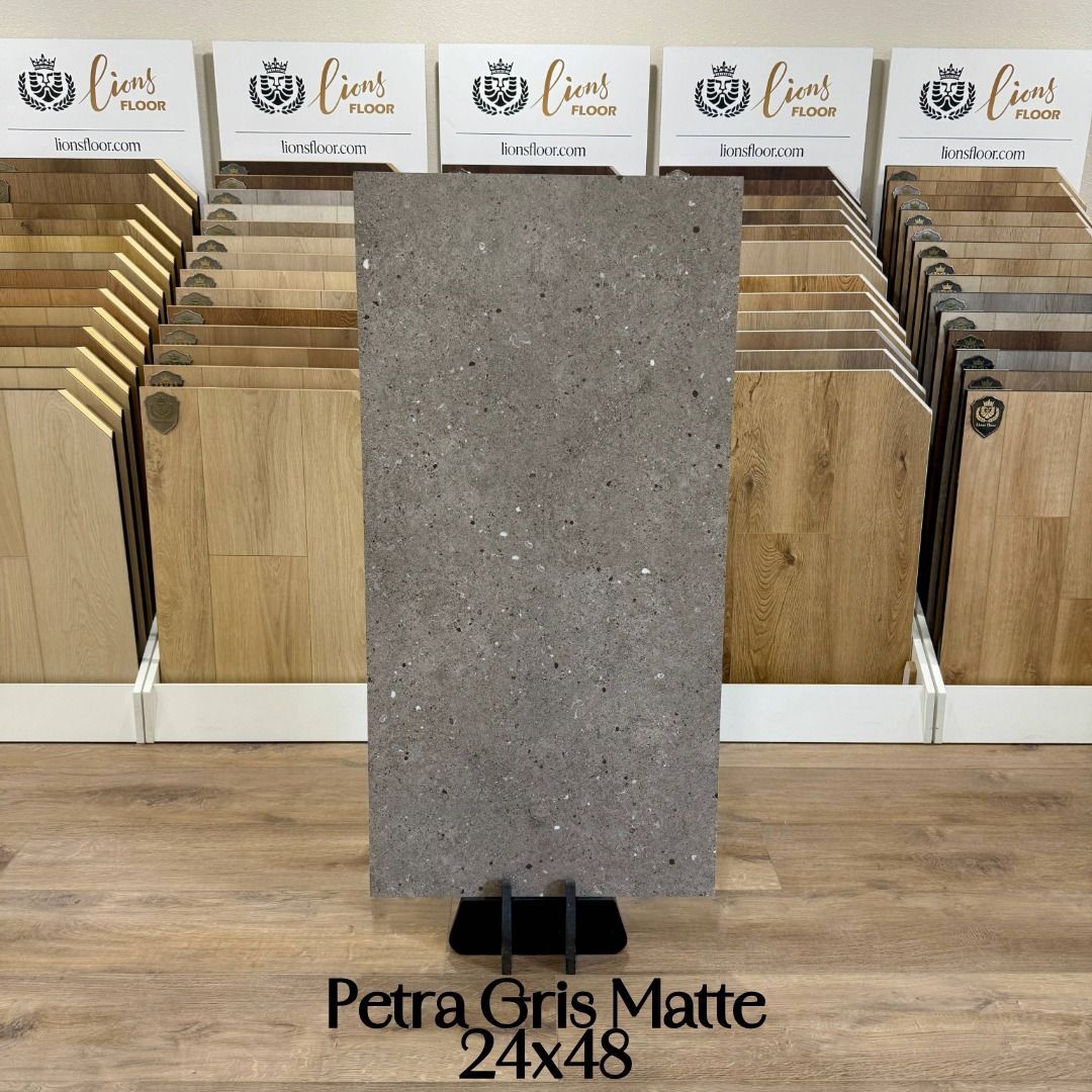 A large, grey, speckled Petra Gris Matte tile displayed on a stand in a flooring showroom with wood planks in the back.