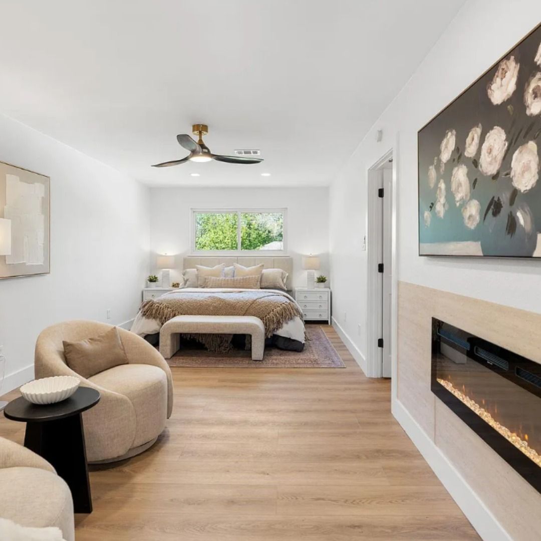 A contemporary master bedroom featuring a large bed, cozy armchair, fireplace, and floral wall art on a light wood floor.