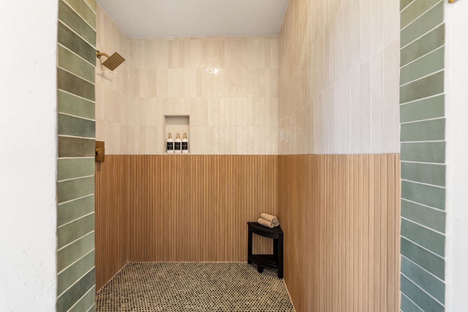 A walk-in shower with vertical sage green tiles on the sides, horizontal wood-look walls, and a pebble floor with a stool.