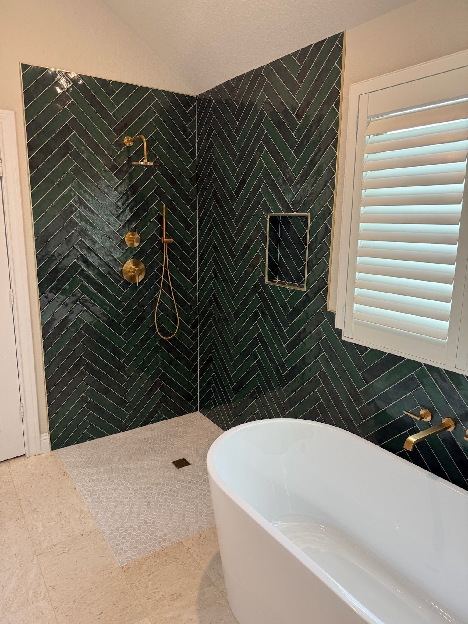 A modern bathroom featuring a white freestanding tub, a walk-in shower with dark green herringbone tile, and gold hardware.