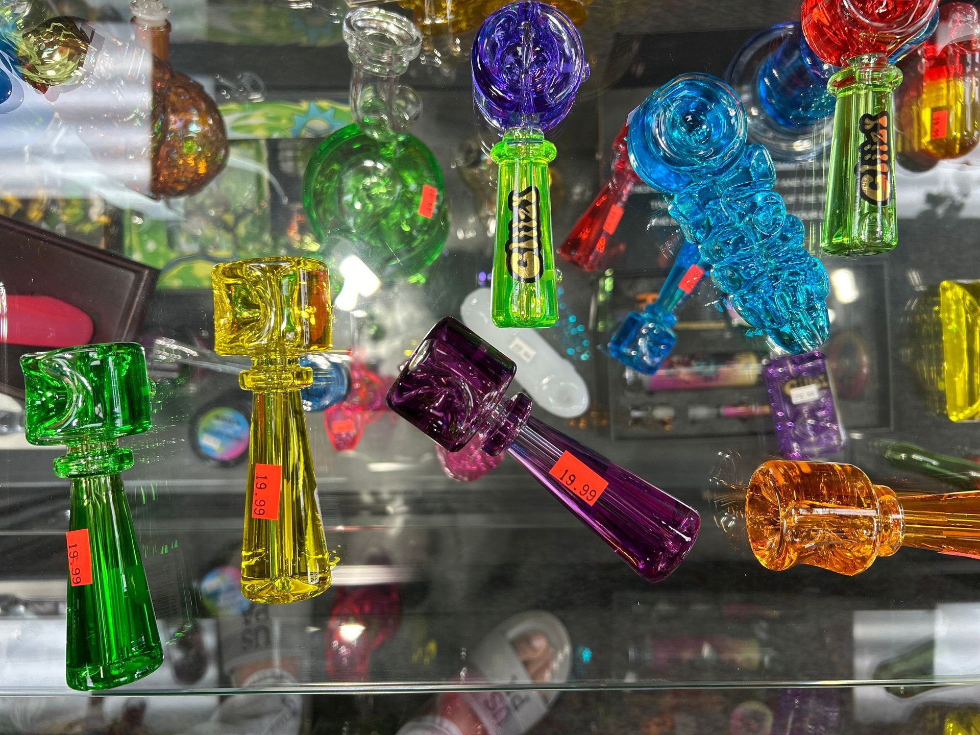 Glass bongs and smoking accessories displayed in a brightly lit glass case, various shapes, and colors.