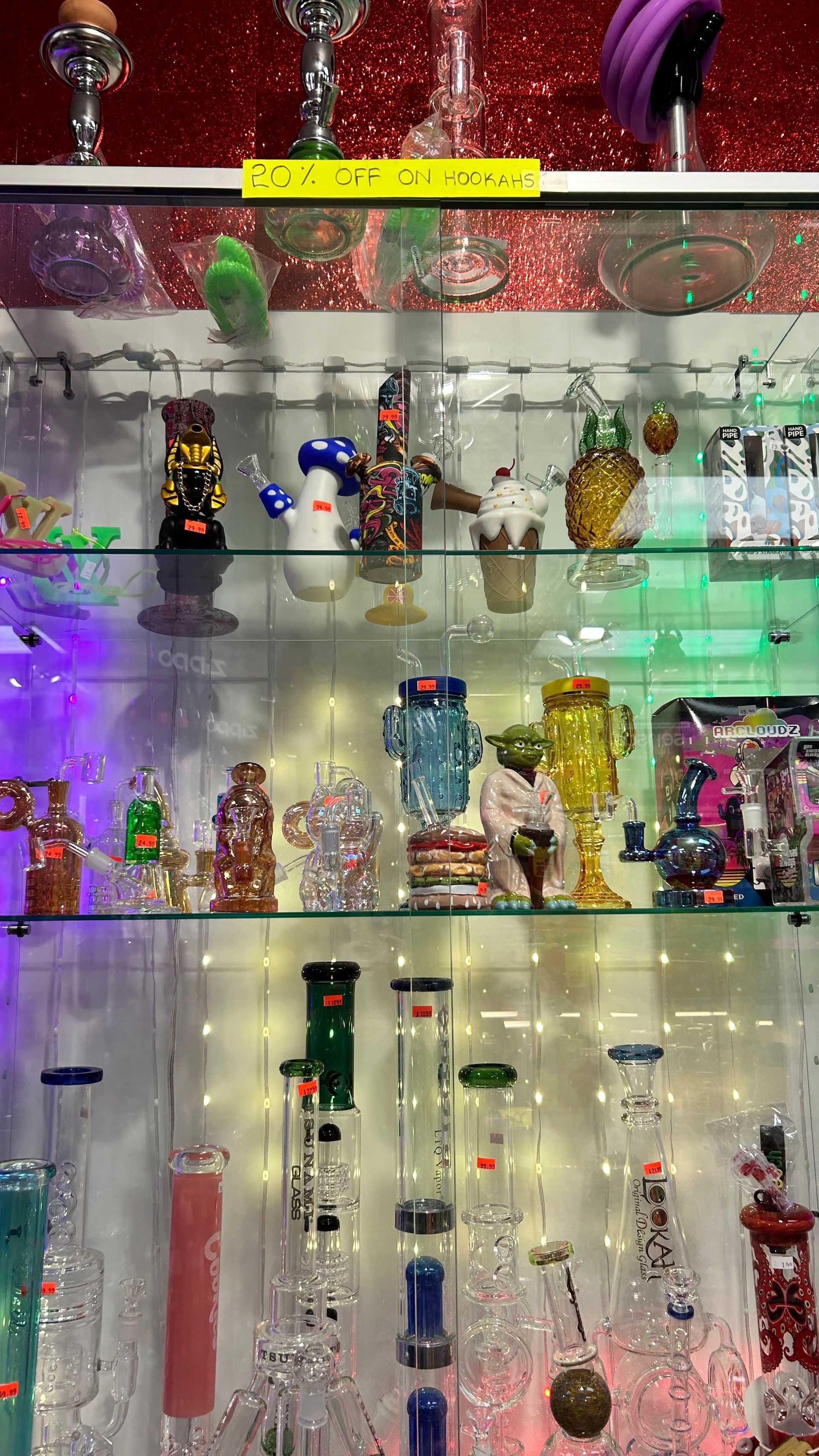Glass display case filled with colorful bongs and decorative smoking pipes, lit by string lights.