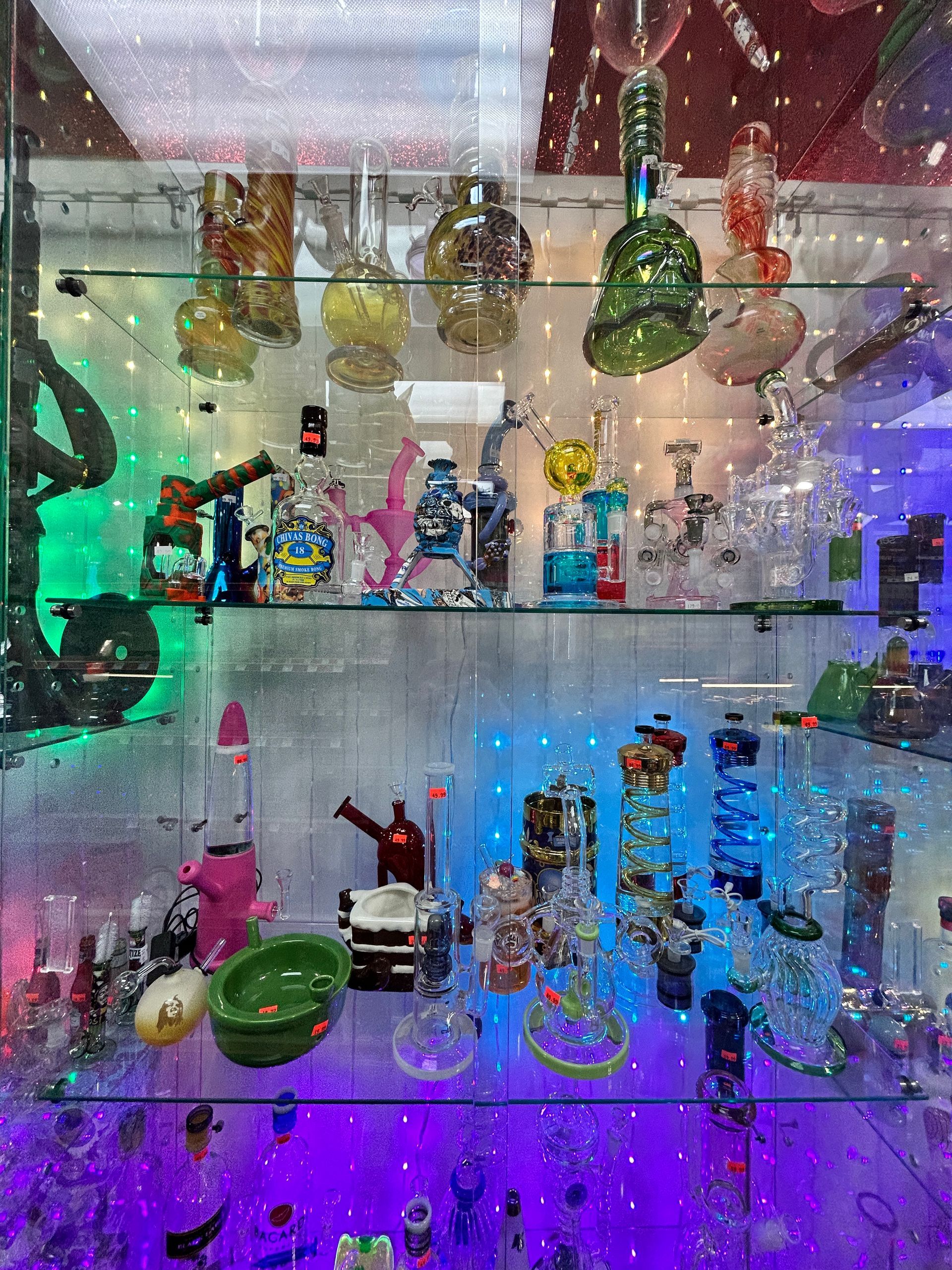 Display case filled with colorful glass bongs and pipes, illuminated by green, blue, and purple lights.