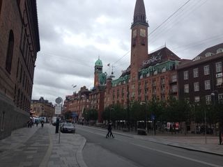 Street view of a building with a clock tower. People walk along the sidewalk and cross the street. Overcast sky.