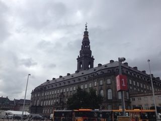 A large, ornate building with a tall spire under a cloudy sky. Buses and streetlights are in the foreground.
