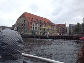 A large, weathered brick building with a red roof sits next to a canal. Water is in the foreground.