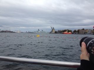 Person holding a camera, taking a photo of a waterway with buoys, ships, and buildings under a cloudy sky.