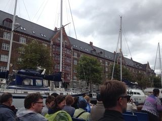Boats and people on a canal. Brick building in the background. Cloudy sky.