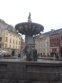 Fountain in a European square; large ornate bronze structure with water spouts; buildings in background.