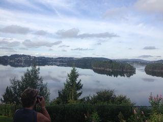 Person photographing a calm lake surrounded by forested hills under a cloudy sky.