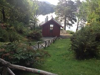 Small, red cabin in a clearing beside a lake, with a wooden fence and greenery.