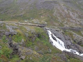 Waterfall cascading over rocks in a mountain landscape, a road with a guardrail runs along the top.