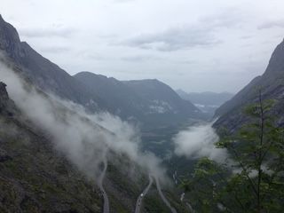Winding road through a mountain valley with low-lying clouds, under a gray, overcast sky.