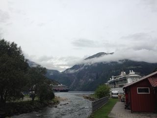 Cloudy mountain fjord scene with cruise ships, red buildings, and a stream.