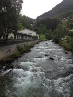 River flowing rapidly through a valley, white water, building along bank, green trees and mountain in the background.