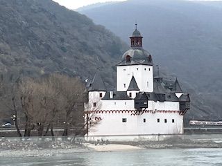 White castle with black trim and a red roof, located on a river, with a mountain in the background.