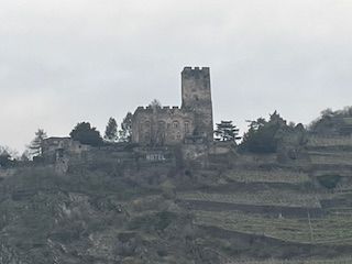 Ruin of a stone castle with a tall tower atop a hillside with terraced vineyards.