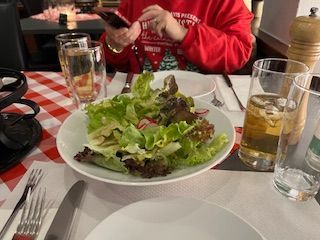 Salad on a plate at a restaurant, with a person in a red sweater using a phone.
