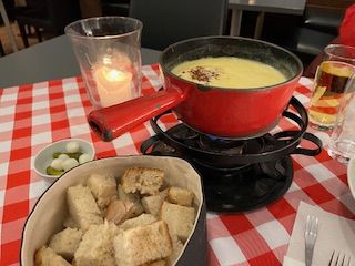 Red fondue pot with cheese, bread, pickles, and candle on a checkered table.