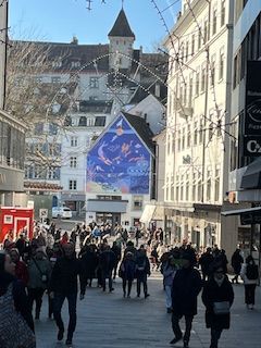 Busy street scene, pedestrians walking buildings and tower in background, mural on building.