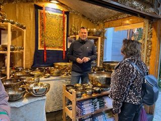 Man in jacket showing customer metal bowls in a shop a blue tapestry is behind him.