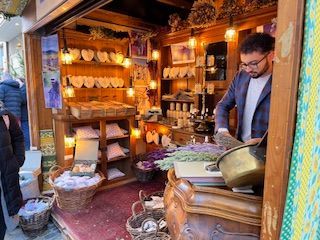 Man working at a shop with lavender products. Wooden shelves display soaps, sachets, and bouquets.