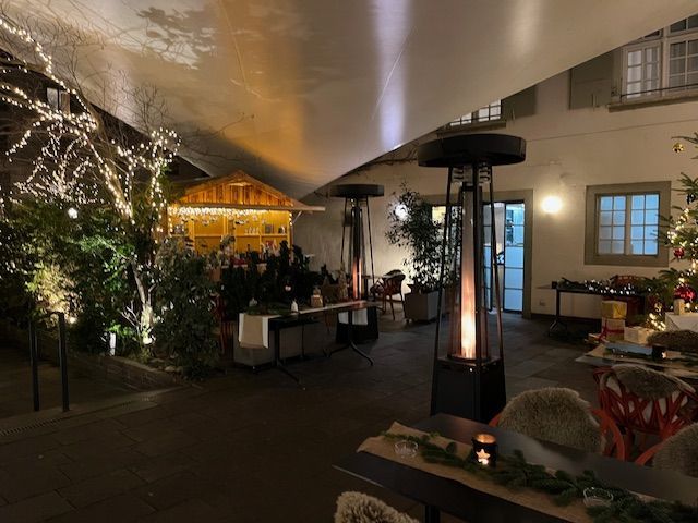 An outdoor Christmas market at night with string lights, heaters, and a wooden hut.
