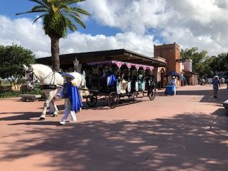 Horse-drawn carriage with passengers travels a brick walkway past buildings, under a palm tree on a sunny day.
