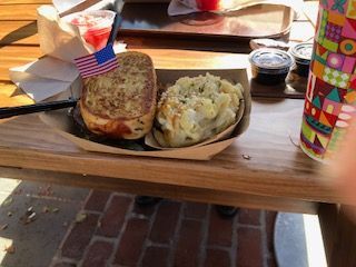 Grilled sandwich and potato casserole in a paper tray, outdoor table, drink with an American flag.