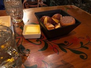 Soft pretzels in a bowl with a side of butter on a floral-decorated table next to two glasses of liquid.