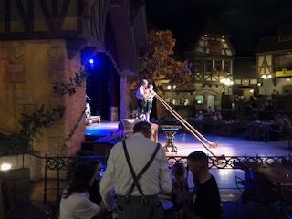 Musicians playing long wooden alphorns on stage at night, with an audience and buildings in the background.
