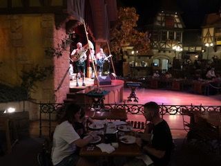People dining, watching a band perform on stage outdoors. Buildings and trees in the background.