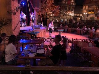 Live band performing on stage at a German restaurant. Audience seated at tables, with a building backdrop.