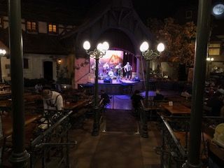 Outdoor restaurant at night with band playing on stage under arched structure, lit by streetlights.