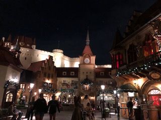 Night scene of a European village square with illuminated buildings and people strolling.
