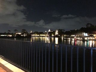 Night view of a waterfront with lights reflecting on the water; buildings and a railing are visible.