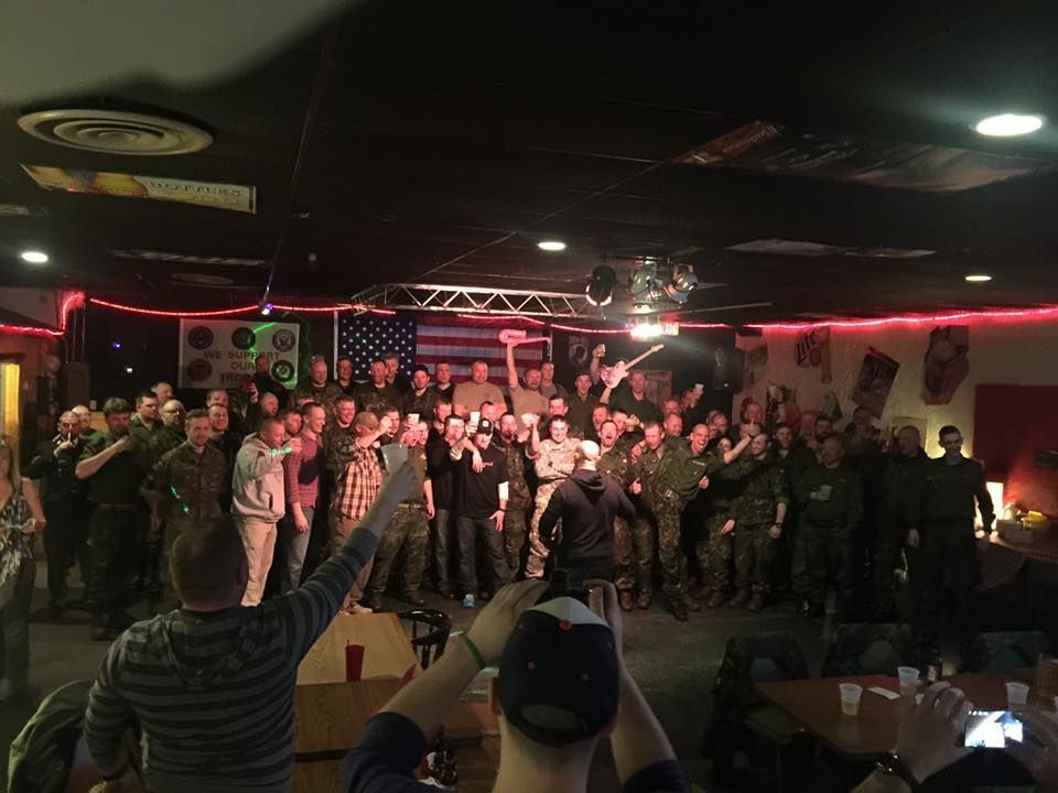 A large group of people are standing in a room with an american flag in the background.