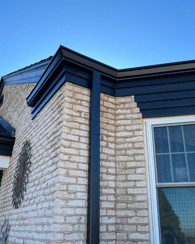 Corner of a brick building with black trim, gutters, and a window against a blue sky.