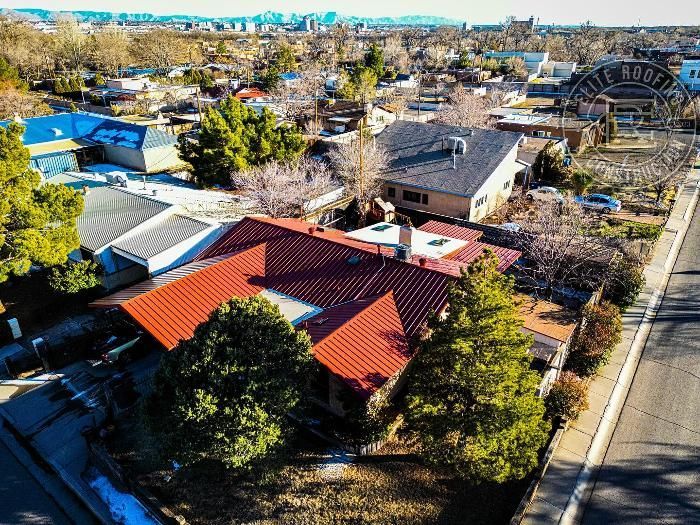 Aerial view of a neighborhood with houses, trees, and a red-tiled roof in the foreground; city skyline visible.