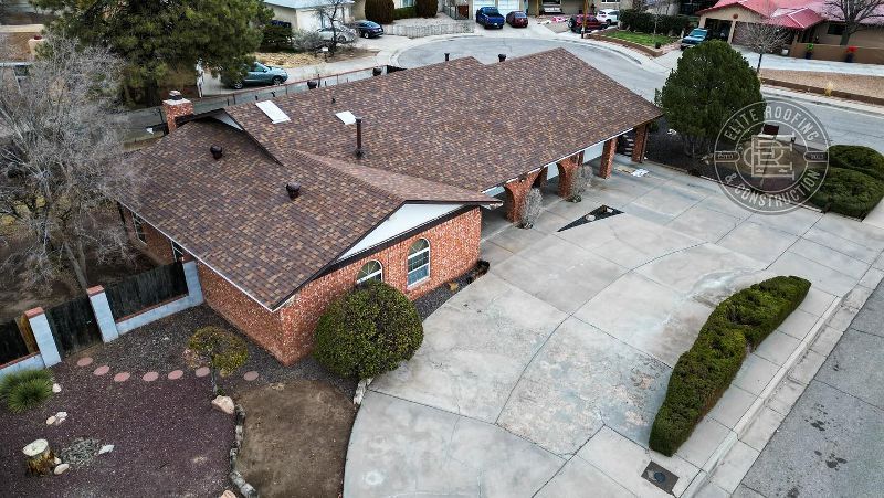 Aerial view of a brick home with a brown roof and driveway, landscaping with greenery and a street.