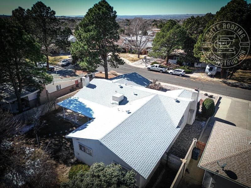 Aerial view of a house with a white roof in a residential area. Trees and cars are in the background.