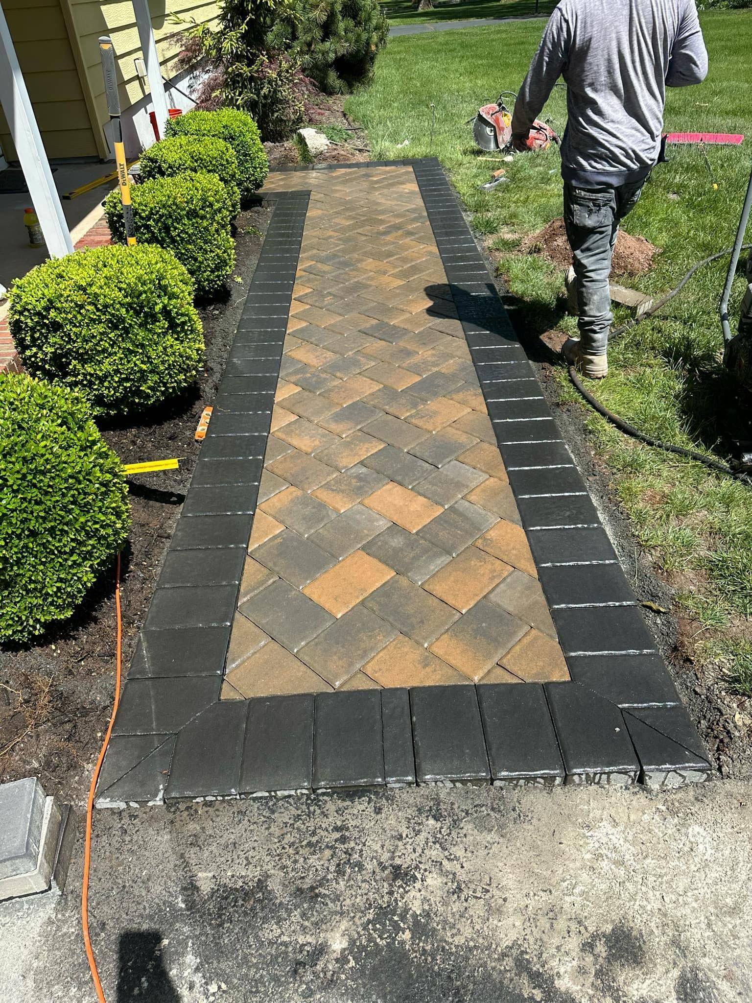 Paver walkway with black border and herringbone pattern, adjacent to shrubs and grass, worker nearby.