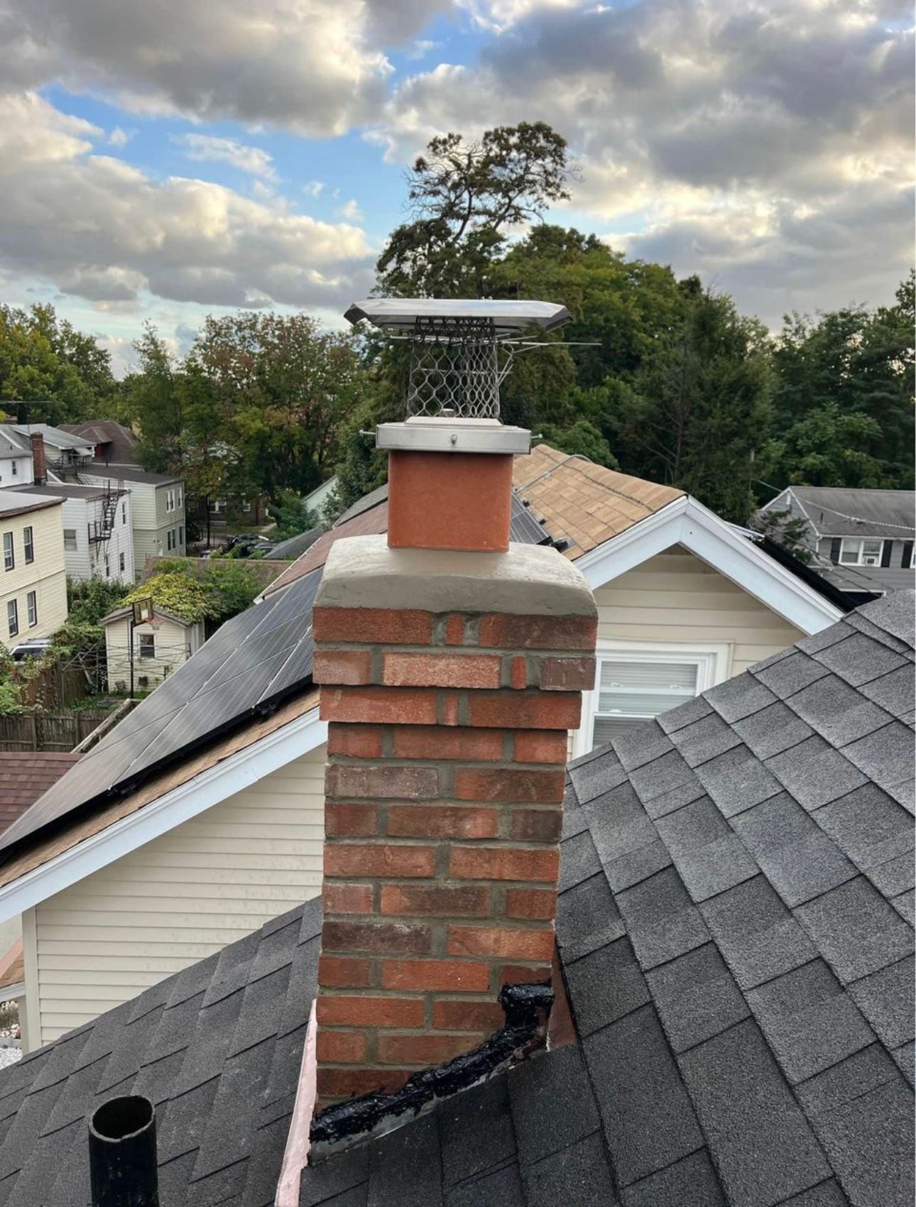 Brick chimney on a rooftop with a metal cap, trees, and houses in the background under a cloudy sky.