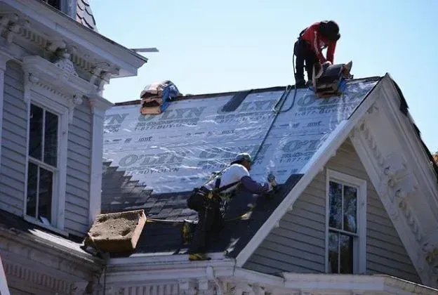 Roofers on a house roof installing new shingles; bright sunlight, clear sky.