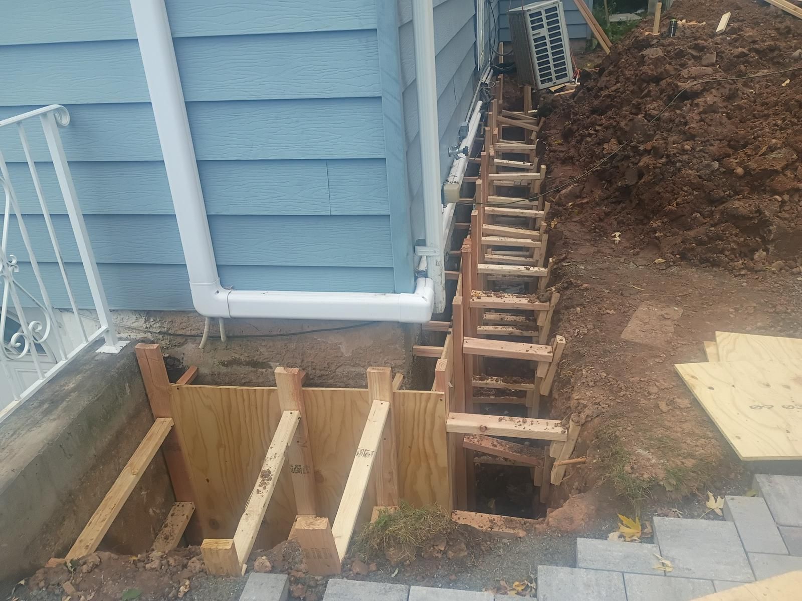 Wooden forms for a concrete structure next to a blue house, with a drainpipe and dirt pile.
