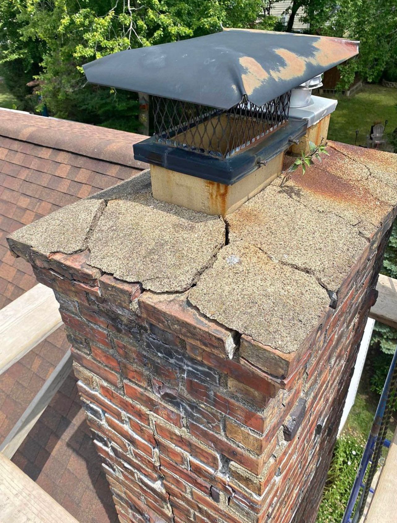 Cracked chimney top with metal cap and brick construction on a roof.