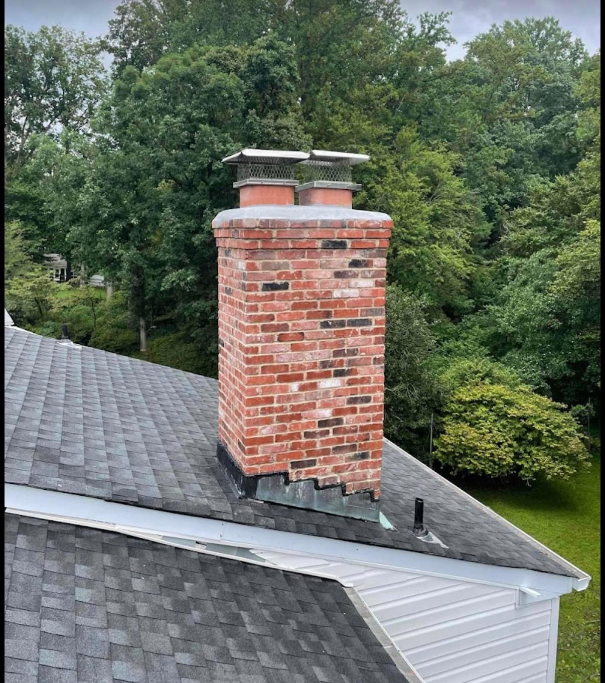 Brick chimney on a dark shingled roof with a backdrop of green trees.